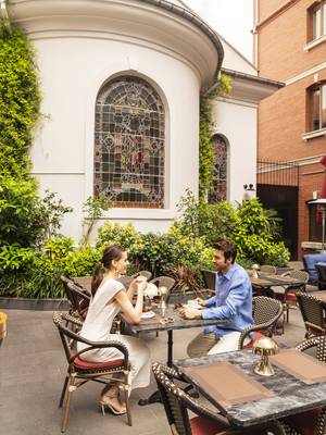 A romantic vertical photo of a couple enjoying coffee at an outdoor table at the Chapelle Restaurant at Rixos Pera Istanbul. They are seated in woven chairs at a small marble table in a courtyard surrounded by lush greenery and a white building with large, ornate stained-glass windows. The woman is wearing a white dress, and the man is wearing a light blue shirt.
