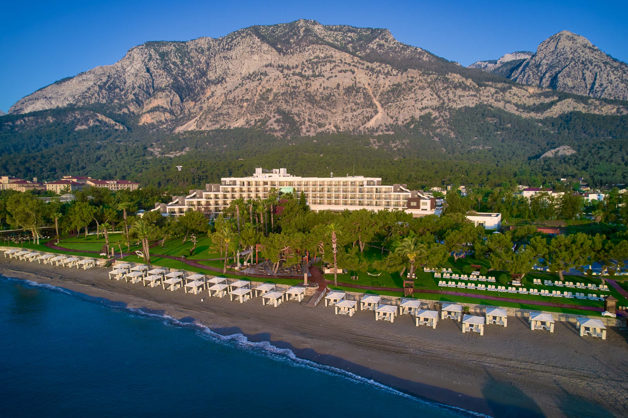 Rixos Beldibi is a beachfront resort with white cabanas along the shore, lush green lawns, and a large hotel building, all set against tall, rugged mountains under a clear blue sky
